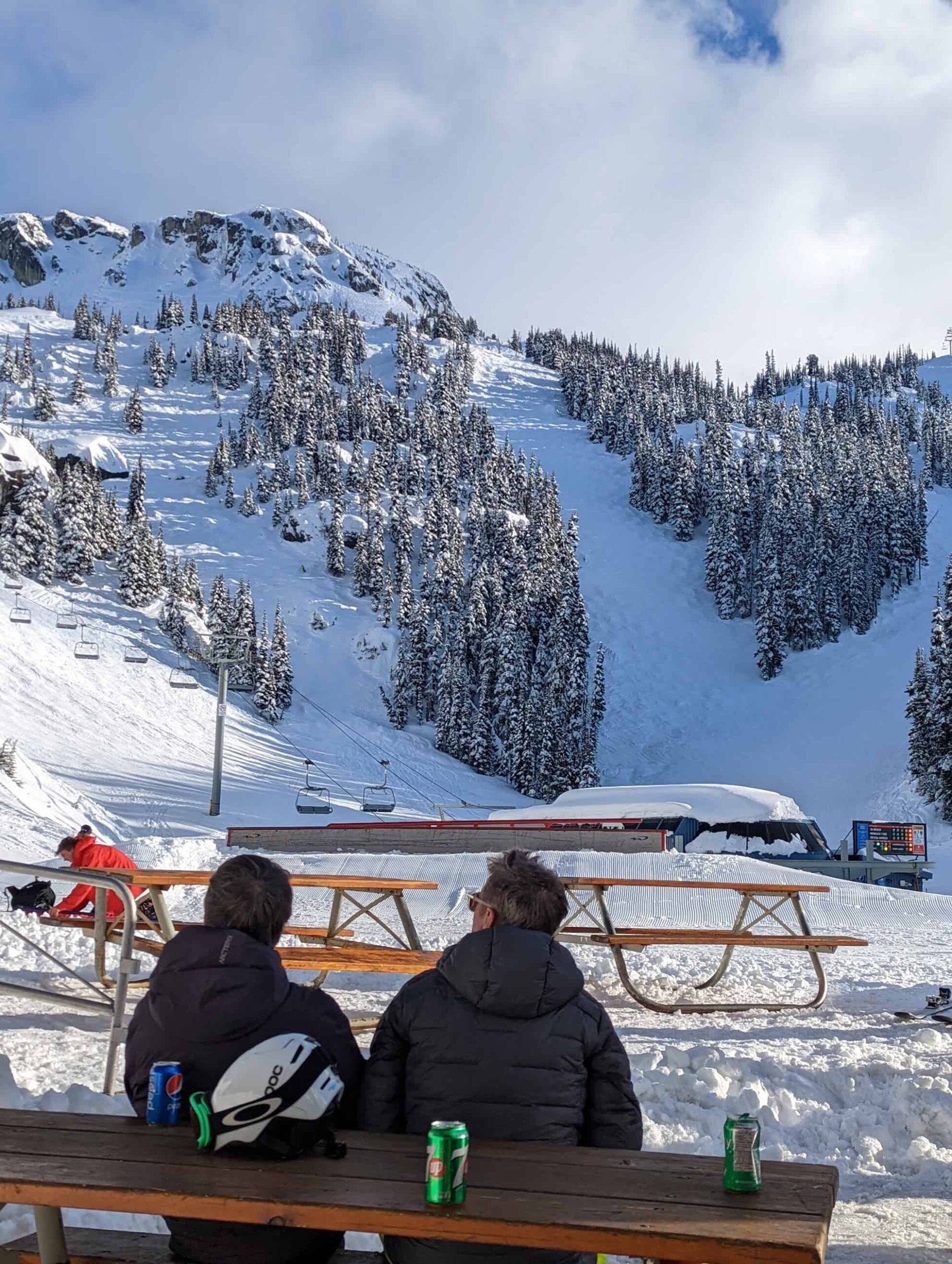 The view from the bottom of Glacier chair lift located on Blackcomb mountain. Two people are sat on a wooden bench. In the background are three ski runs carved in to the side of the mountain. The sky is white and cloudy.