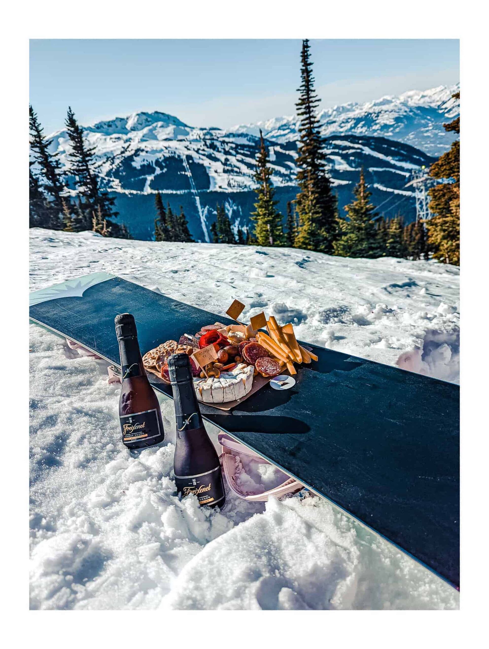 A plate of cheeses sat on top of an upside snowboard. The snowboard is being used as a table. Two small bottles of white wine are being kept cold in the white snow. In the background are snow-capped mountains, pine trees and a pale blue sky.