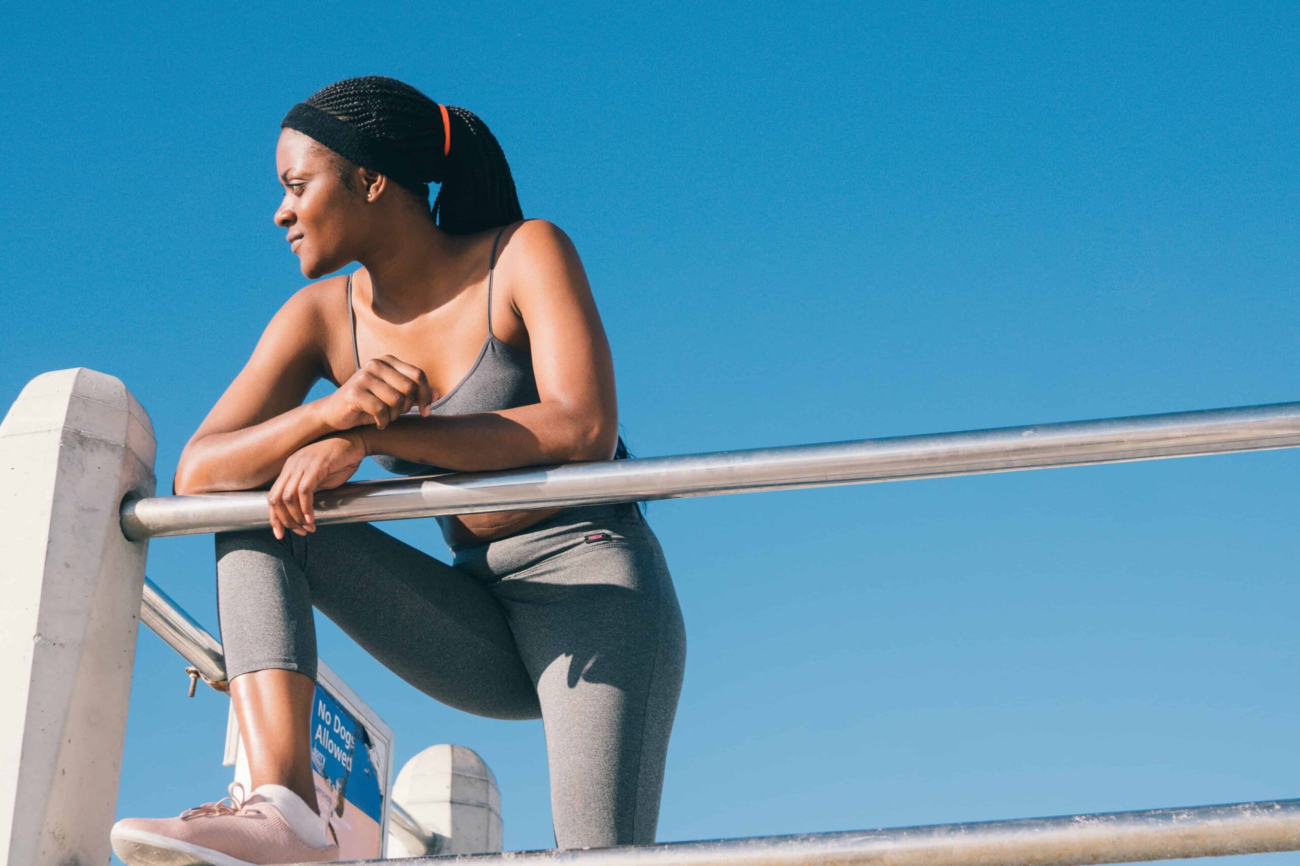 African American girl looking over a fence. She is dressed in exercise clothes and is trying to lose weight. The sky is bright blue behind her.