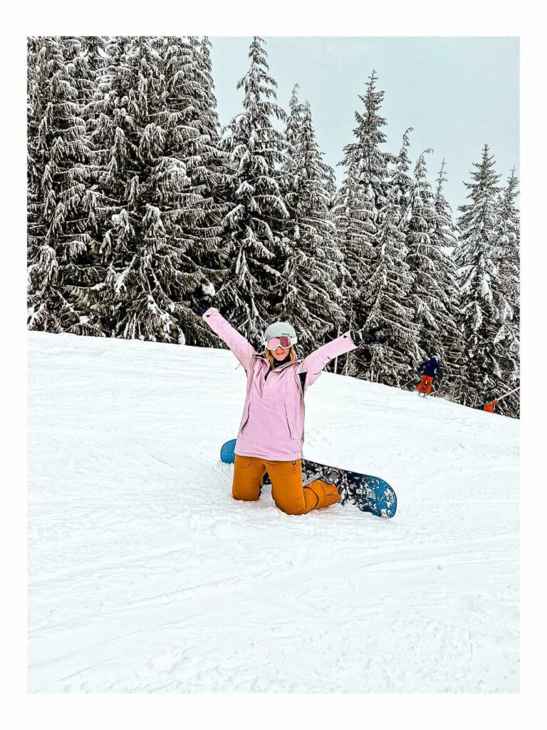 A girl wearing a pink ski jacket, brown ski pants, a grey helmet, pink ski goggles and black gloves, riding a blue snowboard on Whistler mountain