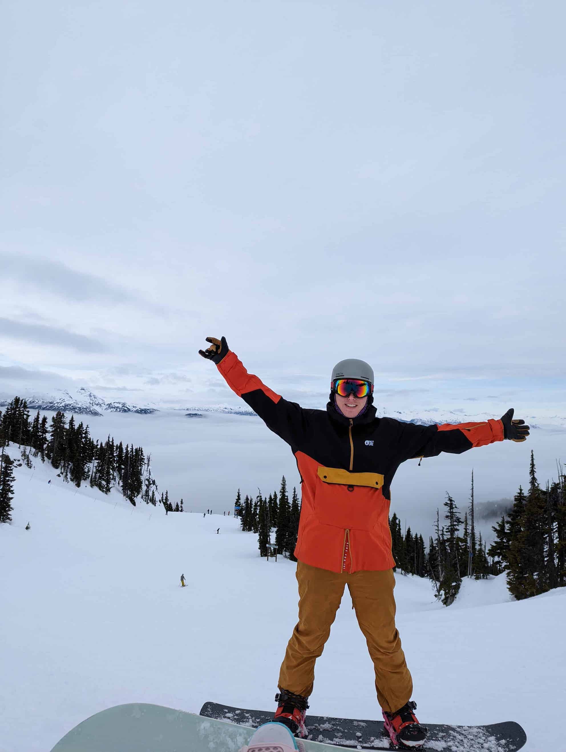 A man in a red and black ski jacket and brown ski pants standing on top of a ski run. The sky is misty and white behind him.