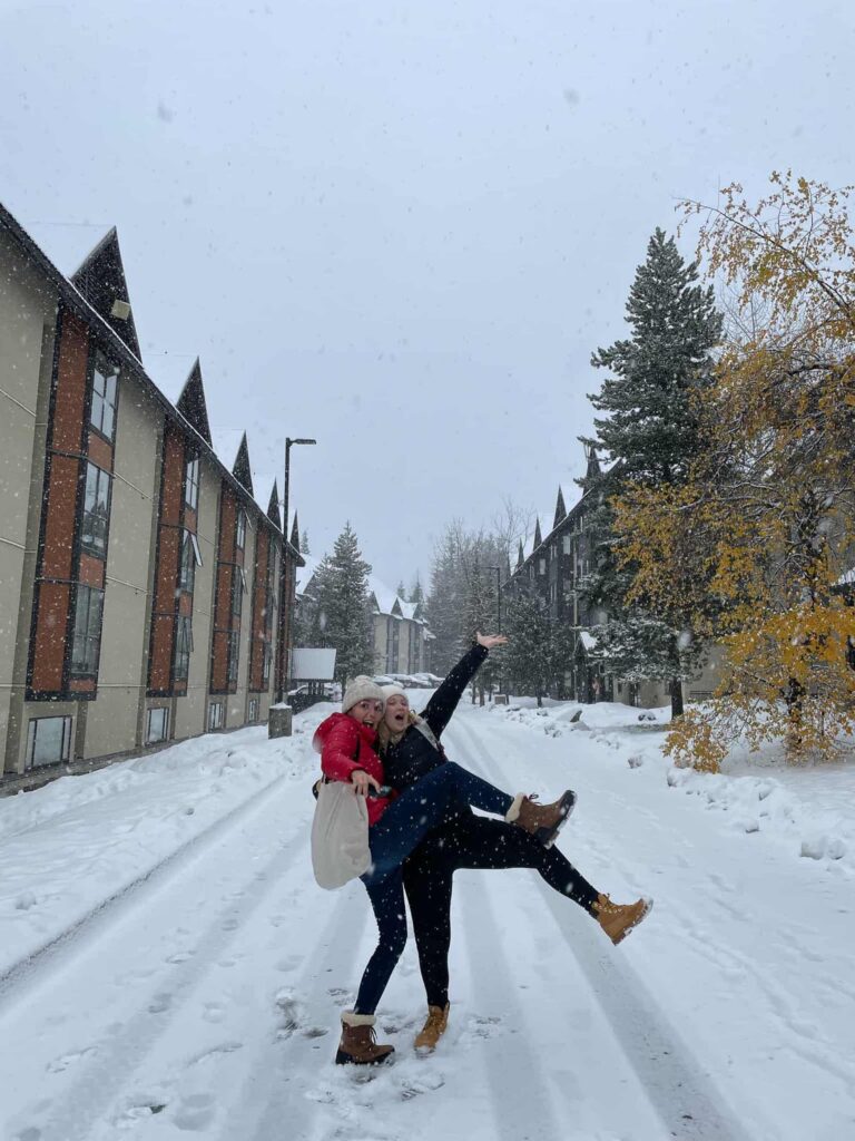 two girls hugging excitedly outside of the Glacier staff housing building which is given to working a ski season in Whistler. The road is blanketed in white snow.