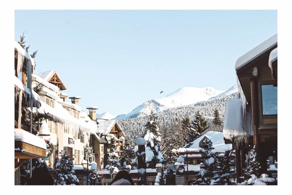 Street photo of Whistler village. the roofs of the shops are blanketed in white snow. Tall snow capped pine tree are in the background against a pale white sky.