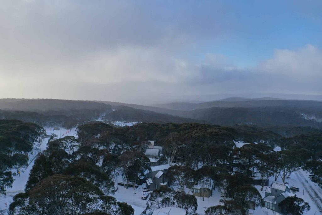 An aerial photo of mount hotham. the sky is dark and misty. the cloud cover is low. tall snow gums poke out of the white ground. 