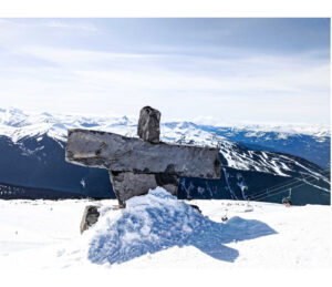 An inunnguat statue on top of the snow capped peaks of Whistler mountain. The sky behind the statue is blue with thin wisps of pale white clouds.