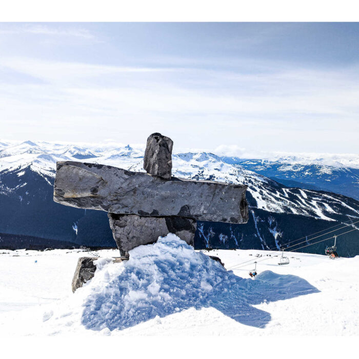 An inunnguat statue on top of the snow capped peaks of Whistler mountain. The sky behind the statue is blue with thin wisps of pale white clouds.