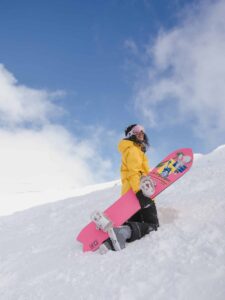 girl wearing ski gear bought in Vancouver not Whistler for a cheaper price. The ski jacket is bright yellow, her pants are black and her snowboard is bright pink.