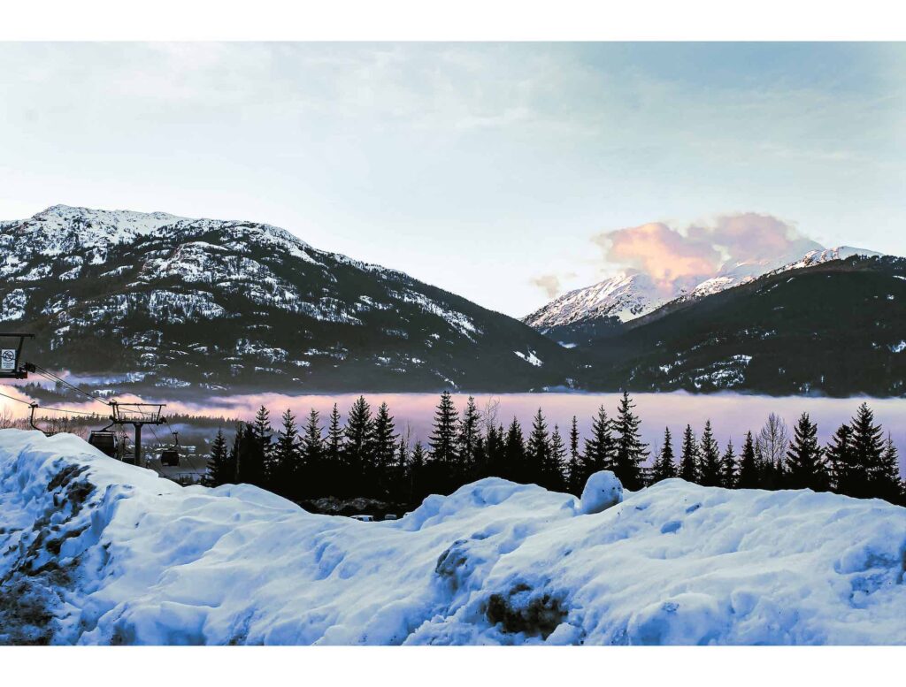 The View from the Glacier staff accommodation car park. White snow has created a tall bank. The Whistler gondola can be seen in the left hand-side of the image. In the Background, two massive mountains are set behind a pink setting sky.